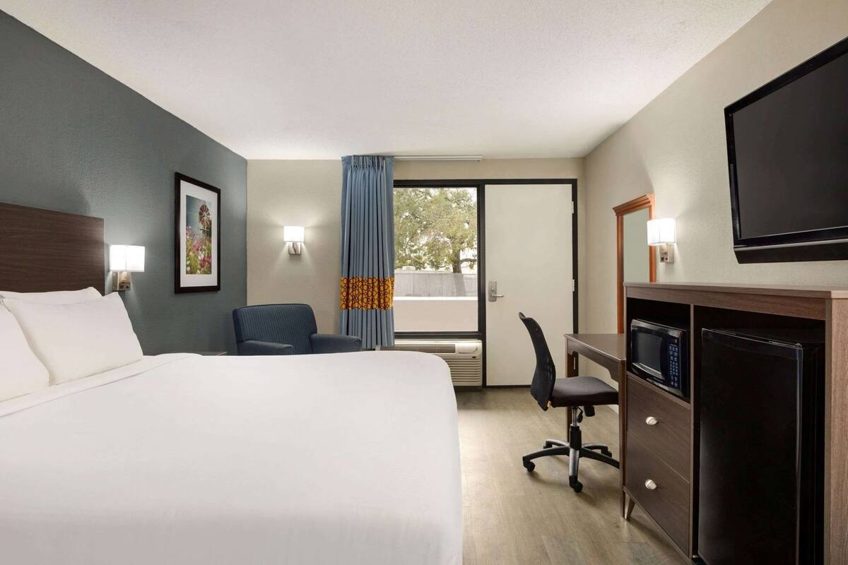 Quarto de hotel com cama, escrivaninha, cadeira, televisão e janela com cortinas.