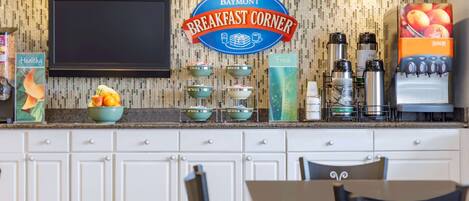 A breakfast area with a Baymont Breakfast Corner sign, a fruit bowl, a juice dispenser, and a coffee machine.