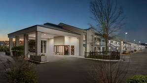 Ein modernes Hotel mit überdachter Eingangspforte, mehreren Stockwerken und einem Parkplatz.