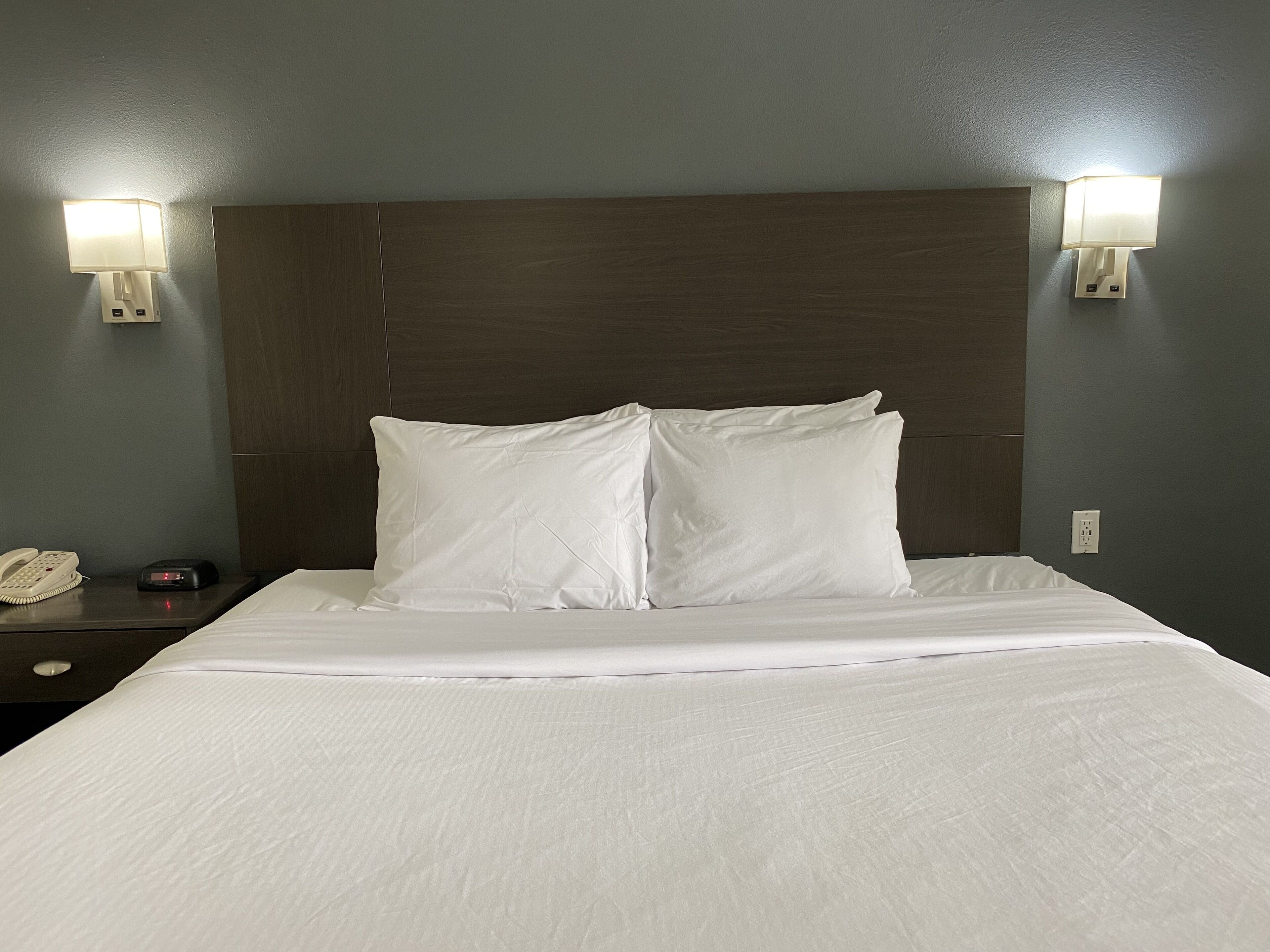 Una habitación de hotel con una cama grande, dos lámparas de noche, una mesita de noche con teléfono y despertador, y un cabecero de madera.