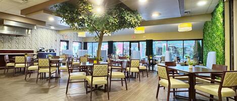 A spacious dining area with wooden tables and chairs, a large tree, and a view of the outdoors through large windows.