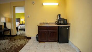 A hotel room with a kitchenette, including a microwave, coffee maker, and sink. There is a bed with a nightstand and lamp in the background.