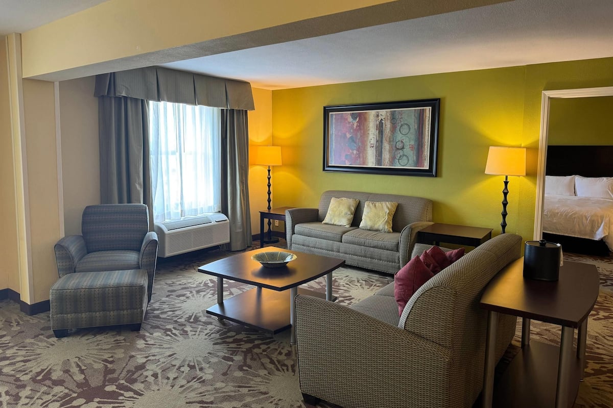 A hotel room with a sofa, armchair, and a small table with a lamp. There is a window with curtains and a painting on the wall.