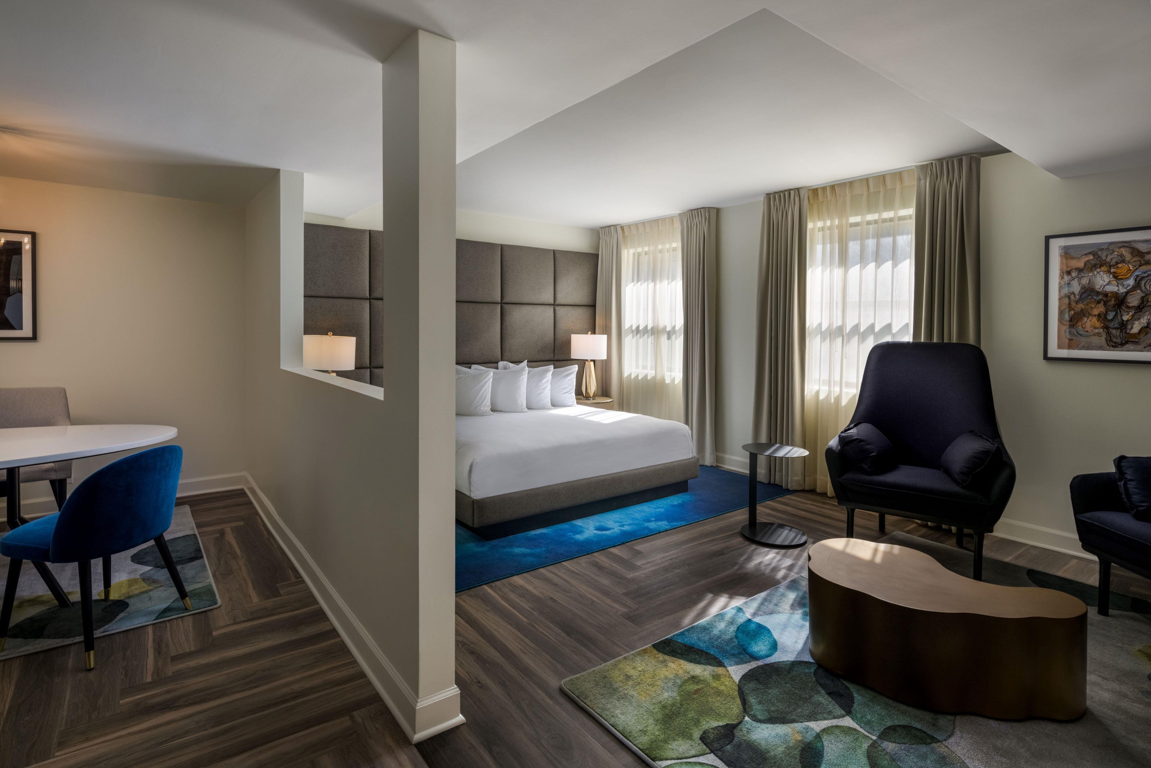 A hotel room with a bed, a desk, a chair, a round coffee table, and a painting on the wall.