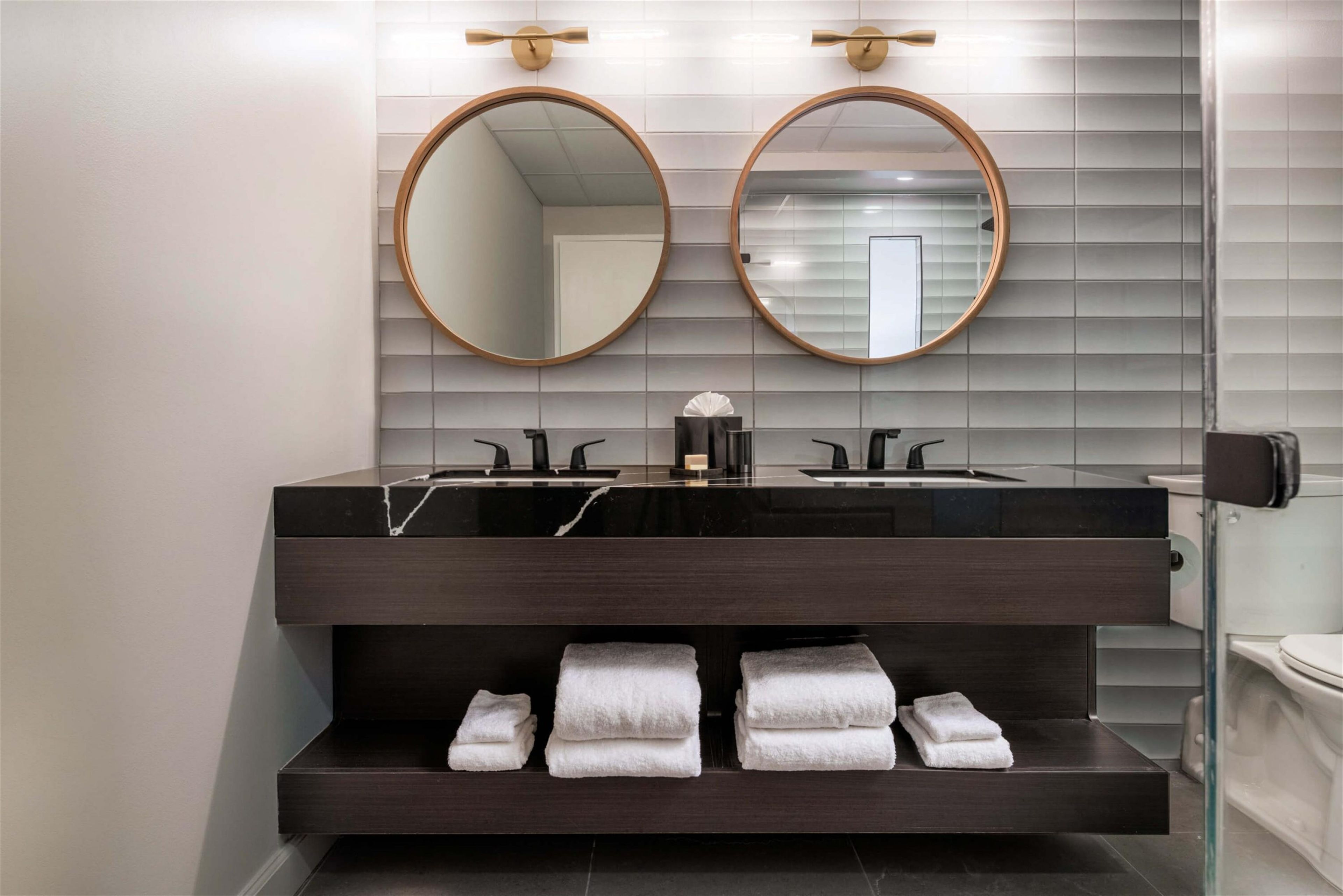 A modern bathroom with a dark marble vanity, two round mirrors, and a towel rack with folded towels.