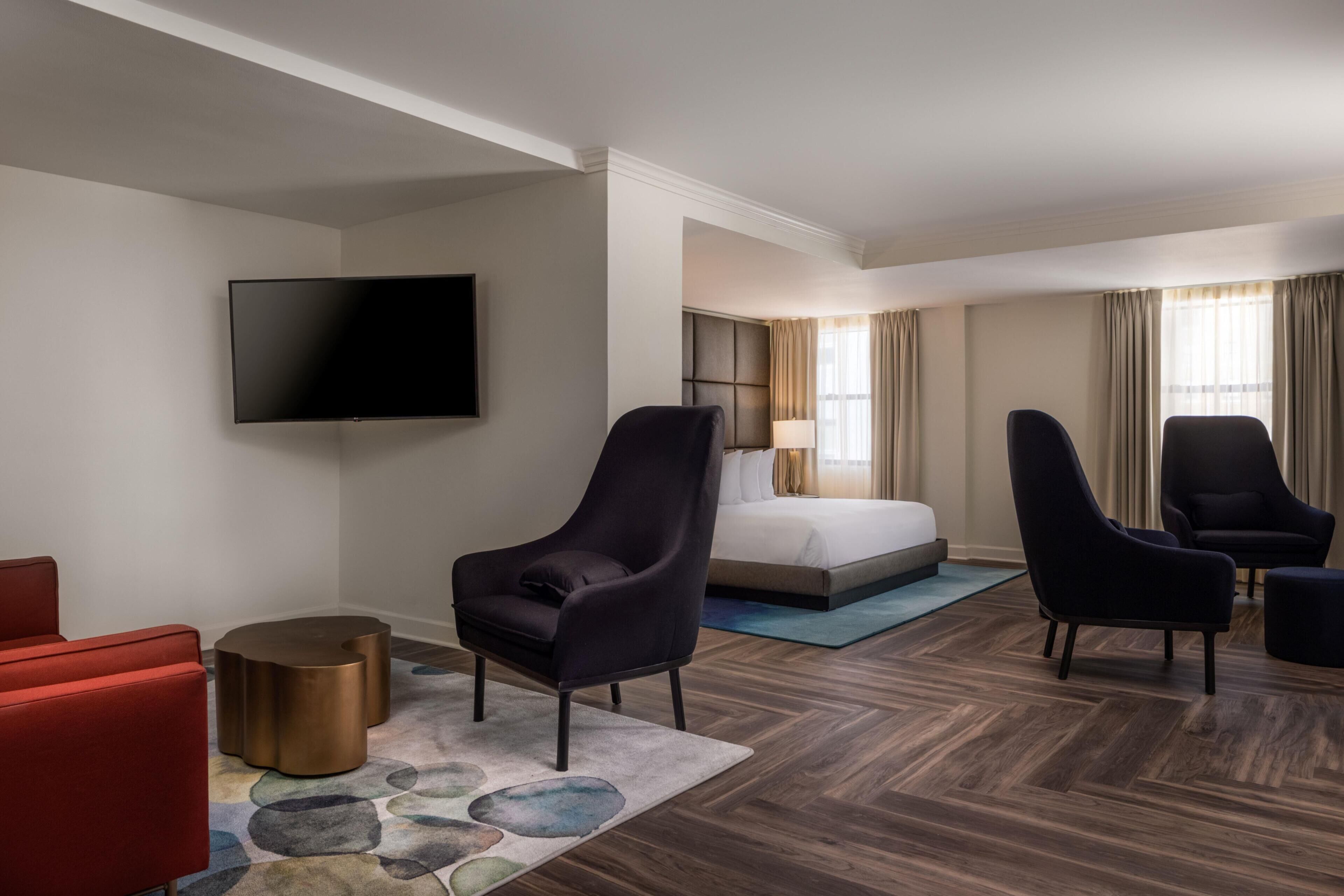 A hotel room with a bed, two armchairs, a red sofa, a flat-screen TV, and a patterned rug.