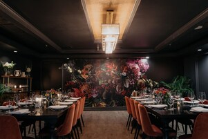 Un restaurant avec une longue table dressée pour le dîner, une fresque florale sur le mur et un lustre au plafond.