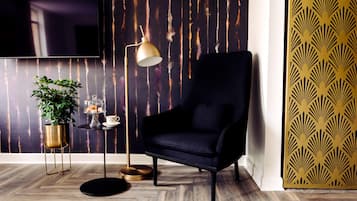 A modern living room with a dark armchair, a floor lamp, a small table with a plant and a decorative item, and a patterned wall.