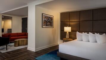 Quarto de hotel com uma cama grande, mesa de cabeceira com abajur, sofá vermelho, mesa de centro e um quadro na parede.