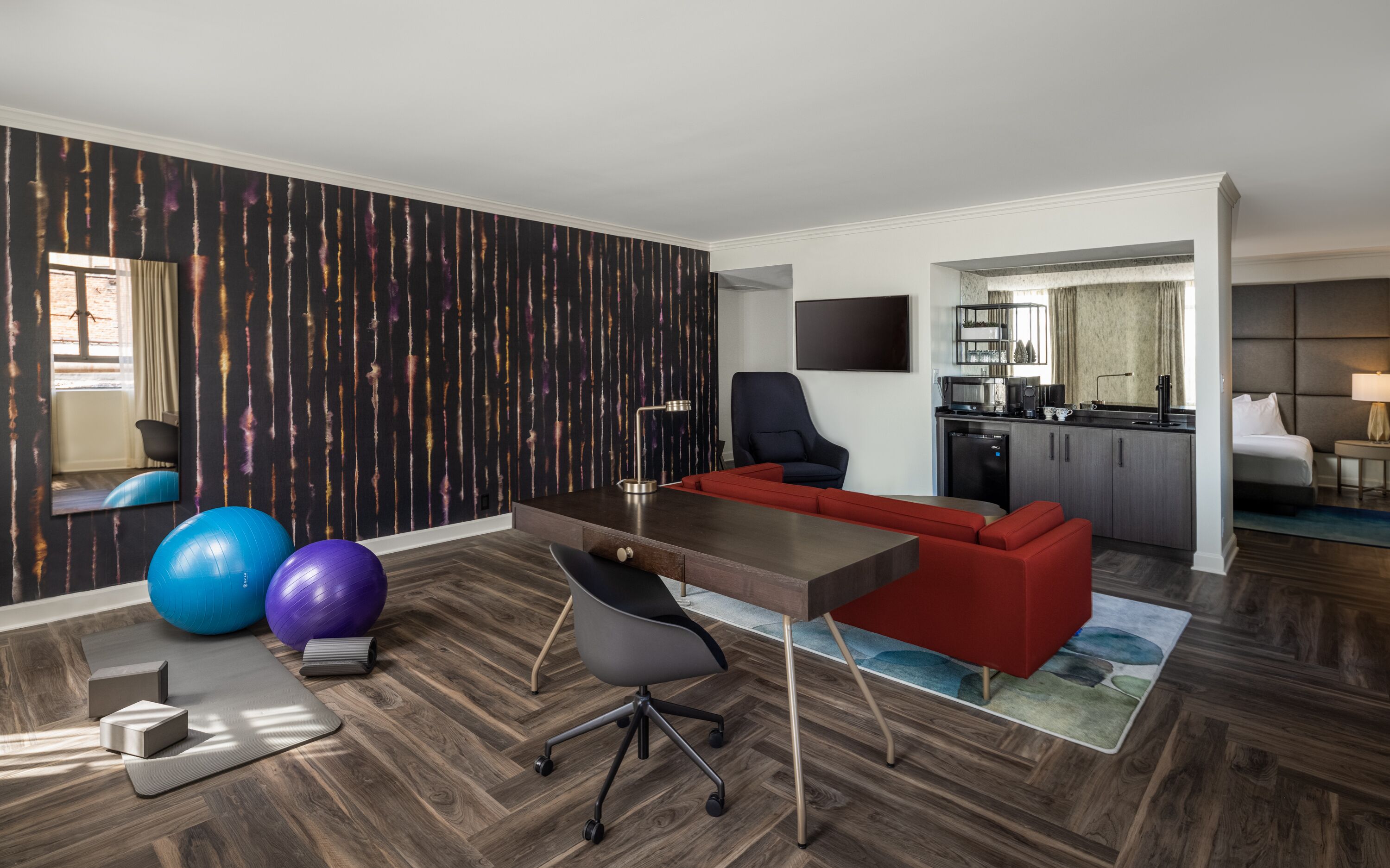 Una habitación de hotel moderna con piso de madera, un escritorio, un sofá rojo, una silla de oficina negra y balones de ejercicio.