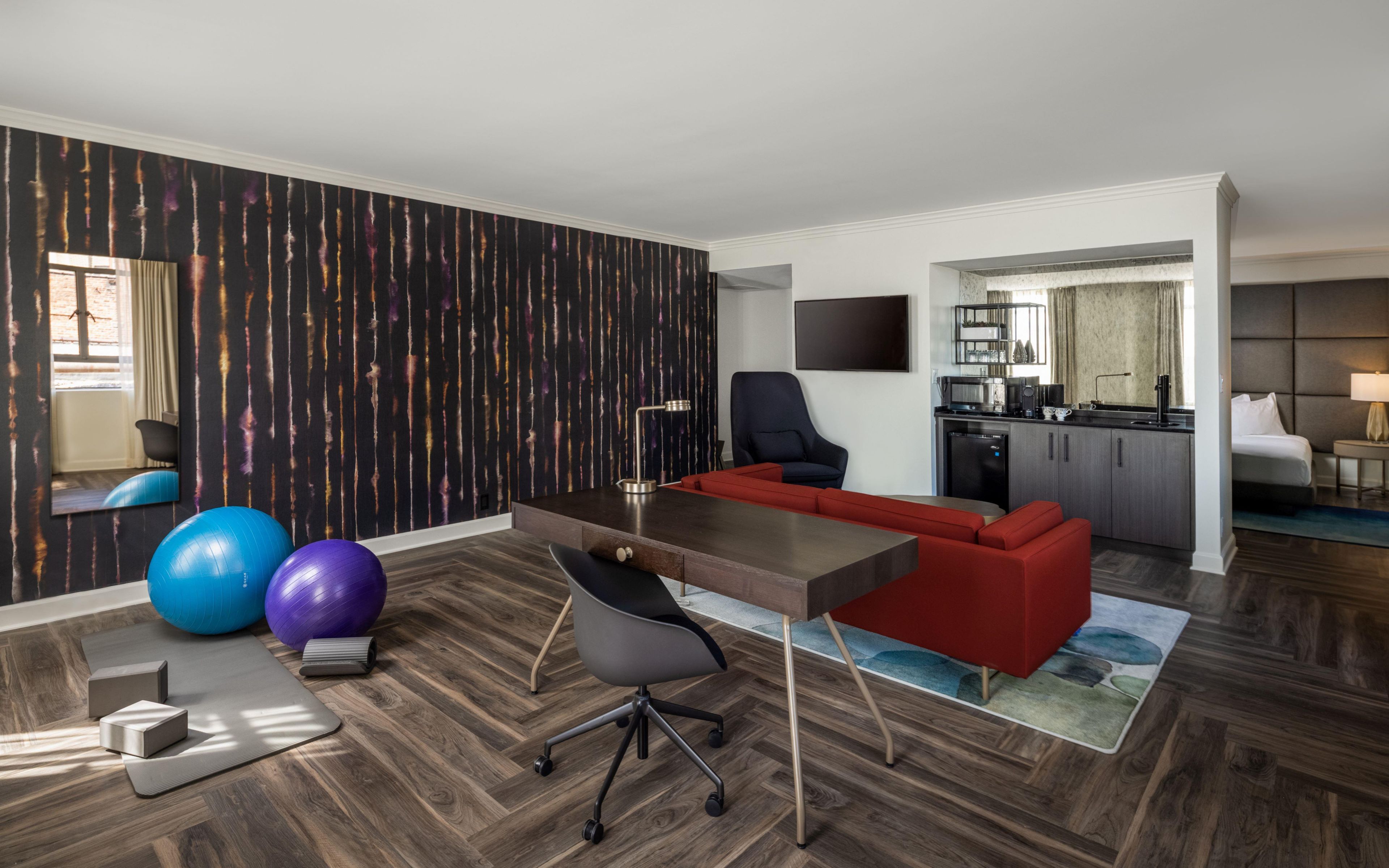 A modern hotel room with a wooden floor, a desk, a red sofa, exercise balls, and a TV.