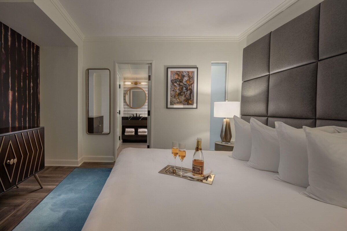 Una habitación de hotel con una cama grande, una mesita de noche con lámpara, un espejo, un cuadro y un minibar.