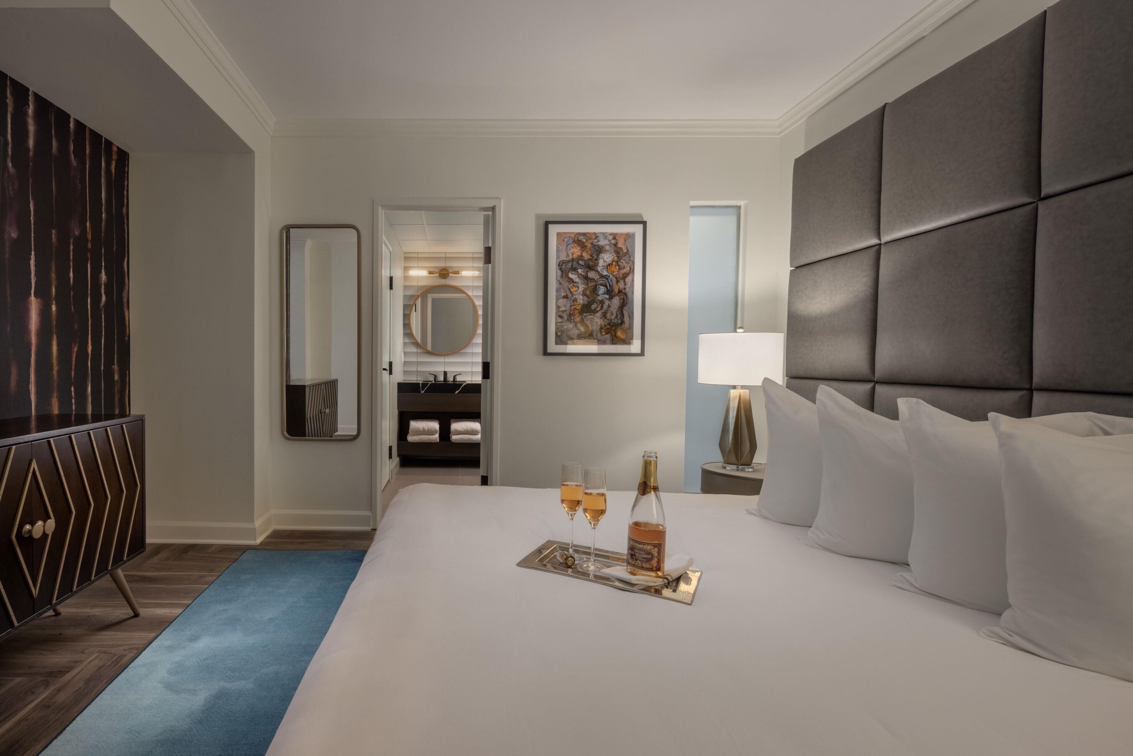 A hotel room with a large bed, a bedside table with a lamp, a mirror, a painting, and a minibar.