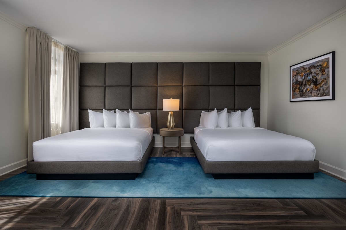 A hotel room with two beds, a large padded headboard, a small bedside table with a lamp, and a blue rug.