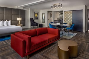 Une chambre d’hôtel avec un lit, un canapé rouge, une petite table ronde, une chaise, une lampe et un coin repas avec une table et des chaises.