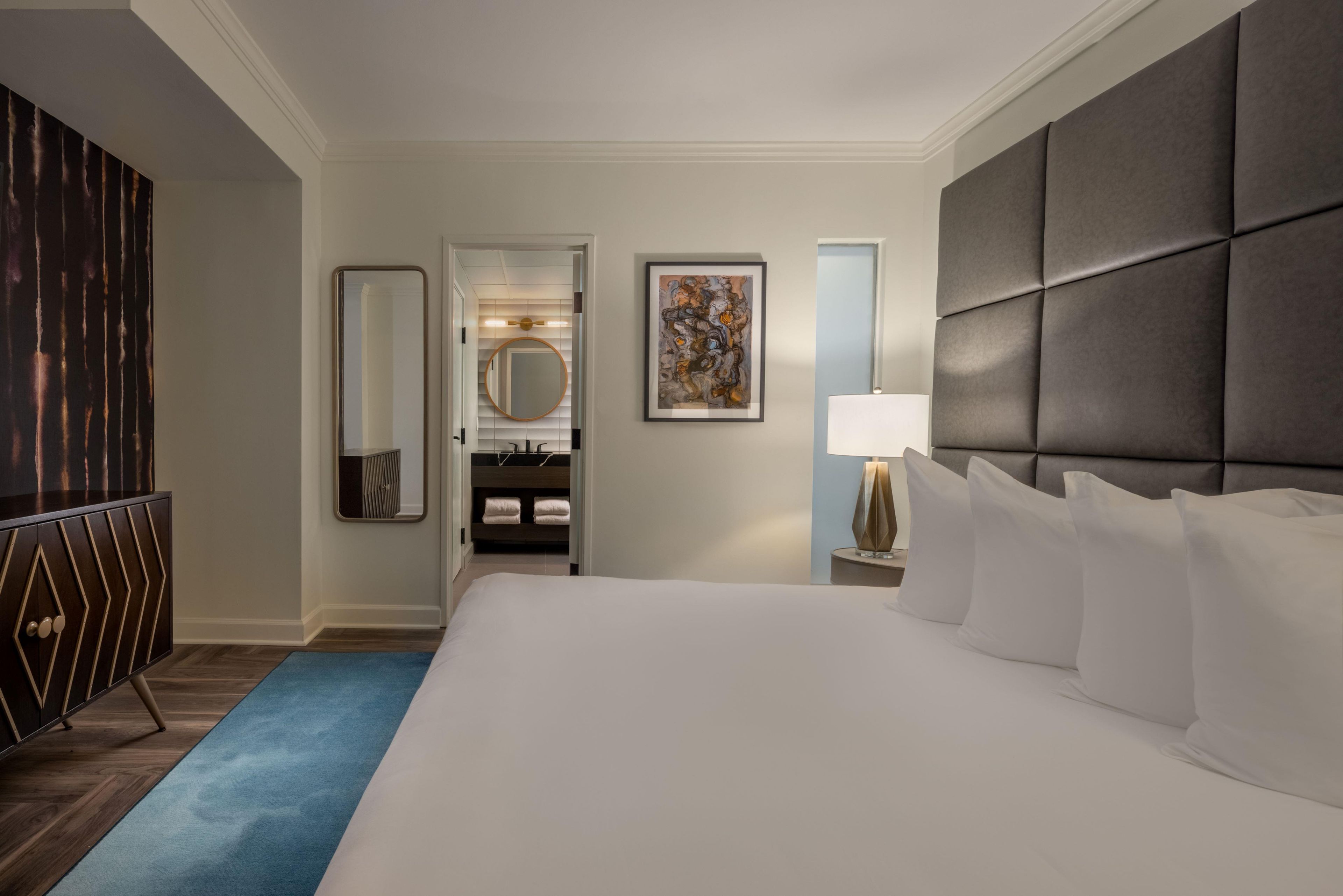 A hotel room with a large bed, a bedside table with a lamp, a wardrobe, a bathroom with a mirror, and a painting on the wall.