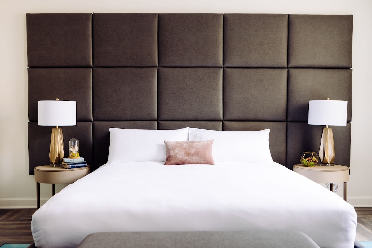 A neatly made bed with a tufted headboard, white linens, and a single decorative pillow. Two bedside tables flank the bed, each with a lamp and a small decorative item.