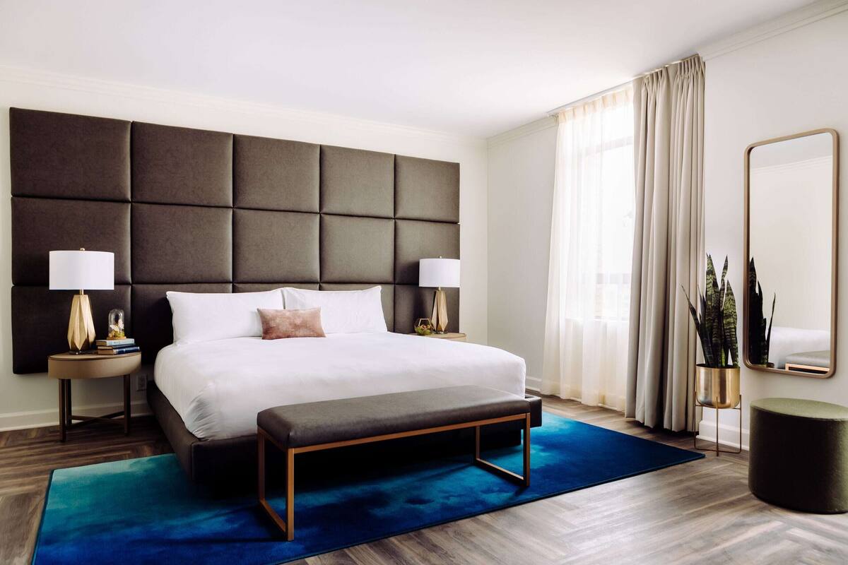 A hotel room with a large bed, two bedside tables with lamps, a bench, a large mirror, and a potted plant.