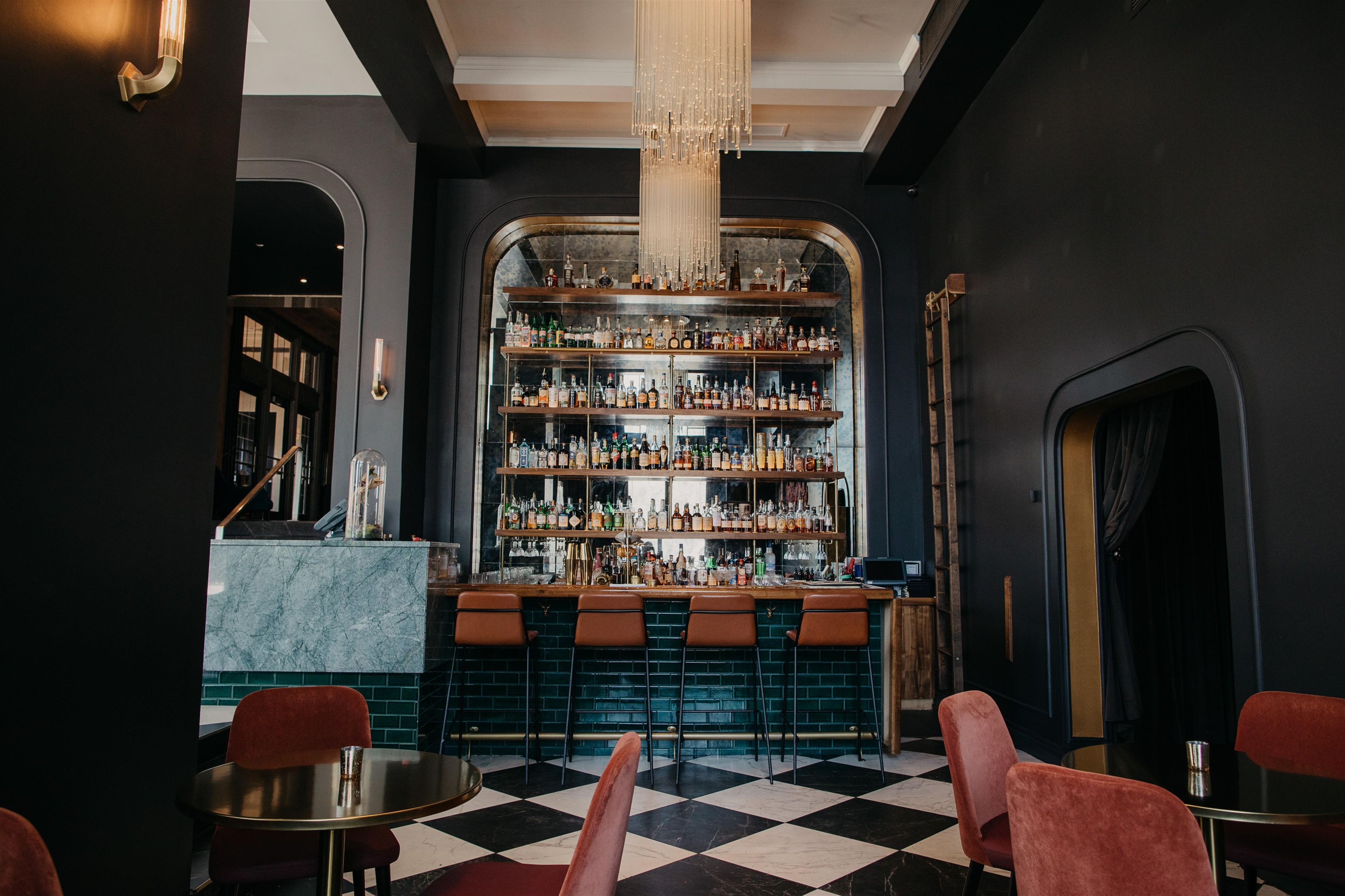 A bar area with a checkered floor, bar stools, and a variety of bottles on shelves.