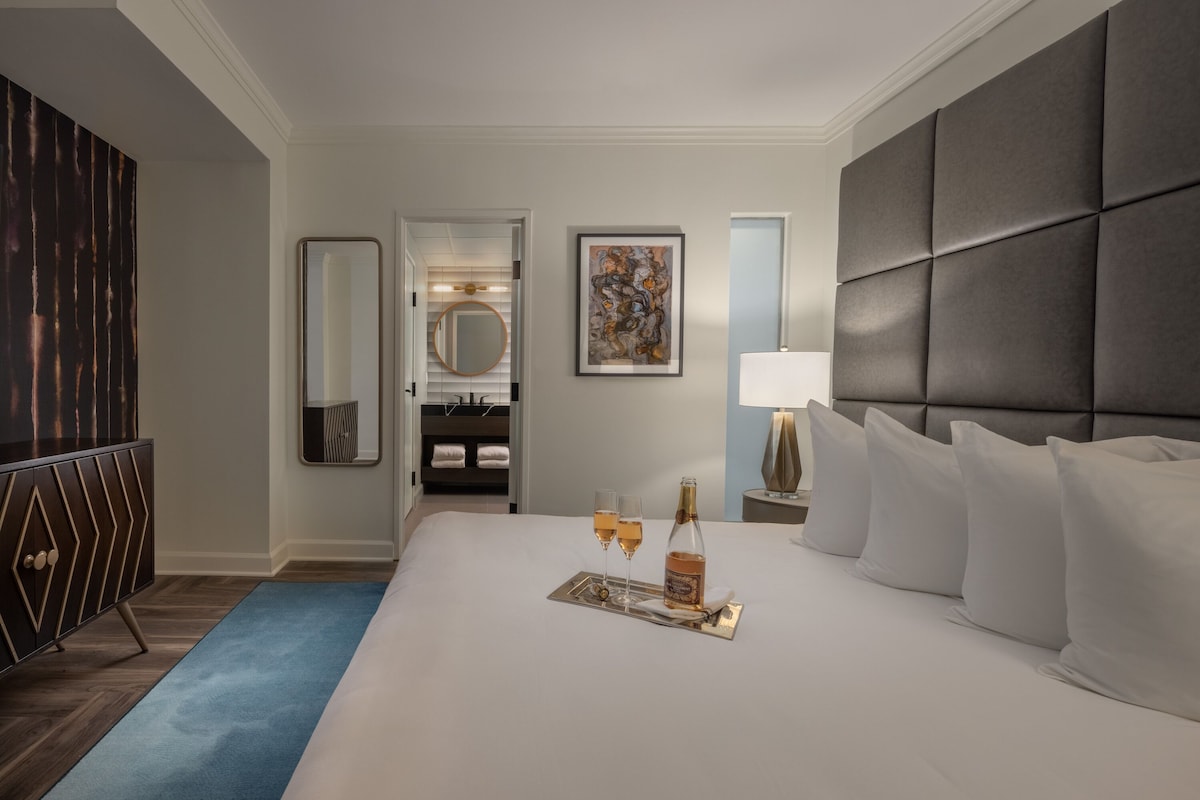A hotel room with a large bed, a dresser, a mirror, a painting, and a bedside table with a lamp.
