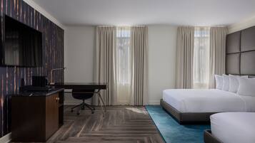 A hotel room with a large bed, a desk with a chair, a flat-screen TV mounted on the wall, and a window with curtains.