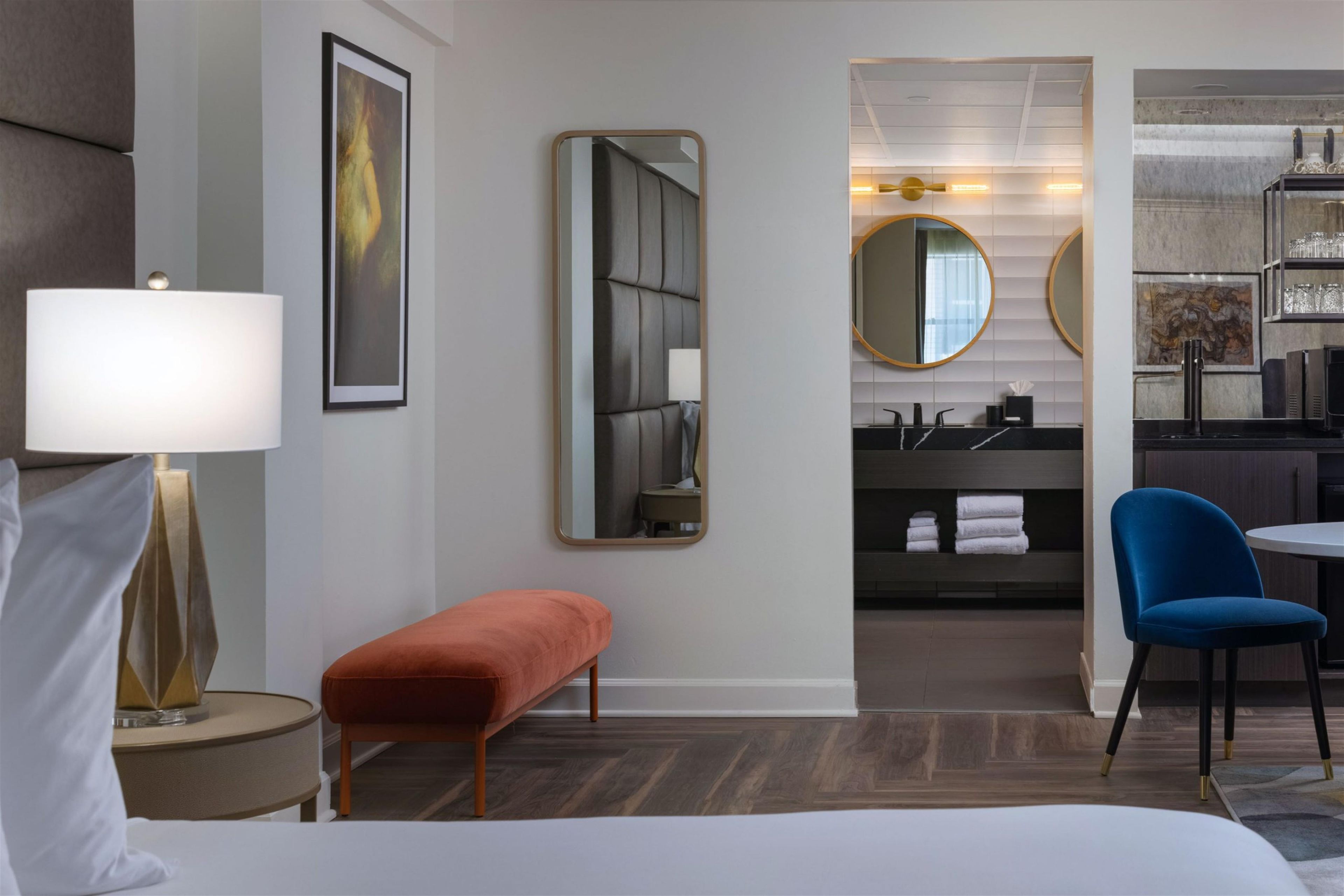 A hotel room with a bed, a bedside lamp, a mirror, a bench, a round table, a blue chair, and a bathroom with a sink and towel rack.