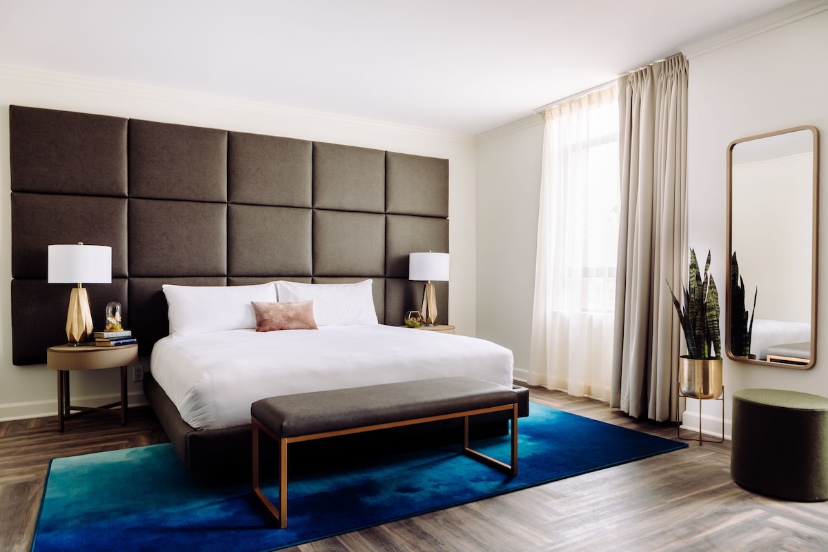 A modern hotel room with a large bed, two bedside tables, a bench, a large mirror, and a potted plant.