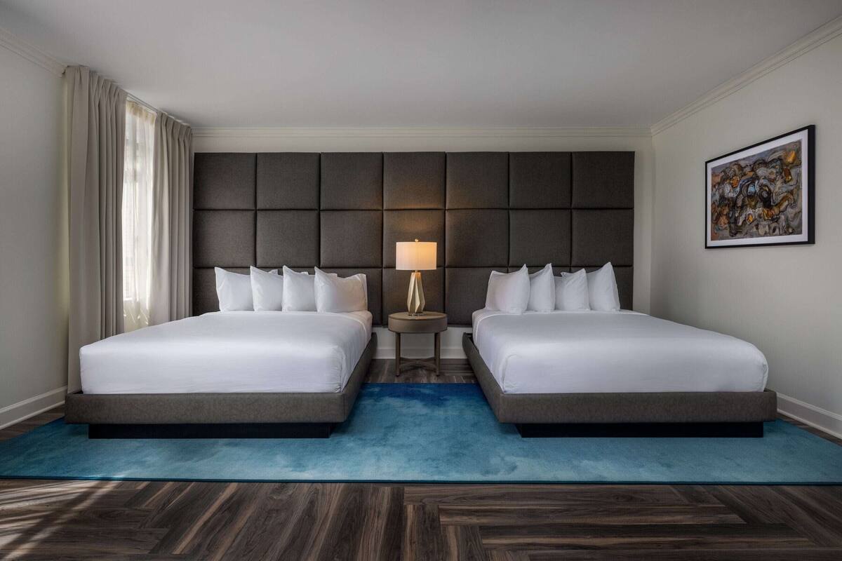 A hotel room with two beds, a large padded headboard, a bedside lamp, a blue rug, and a framed artwork on the wall.