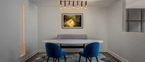 A dining area with a round table, two blue chairs, a modern chandelier, and a framed abstract painting on the wall.