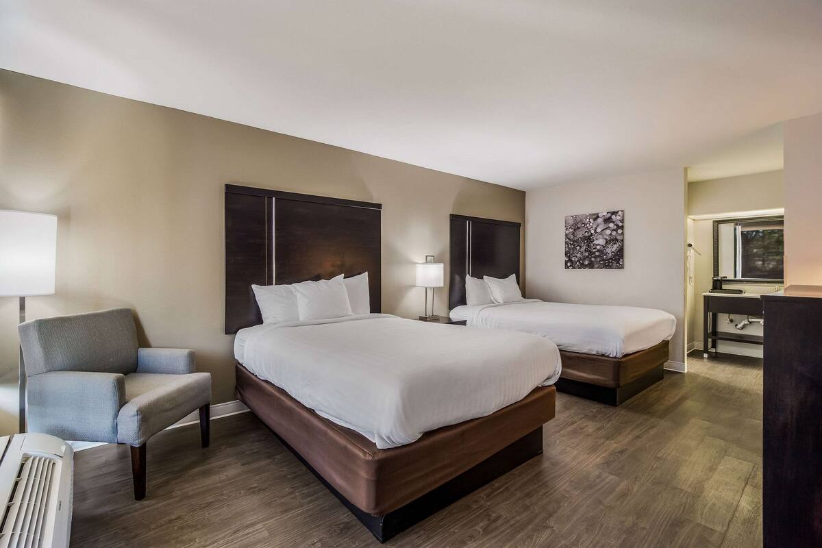 Quarto de hotel com duas camas, uma cadeira, uma escrivaninha e um abajur.