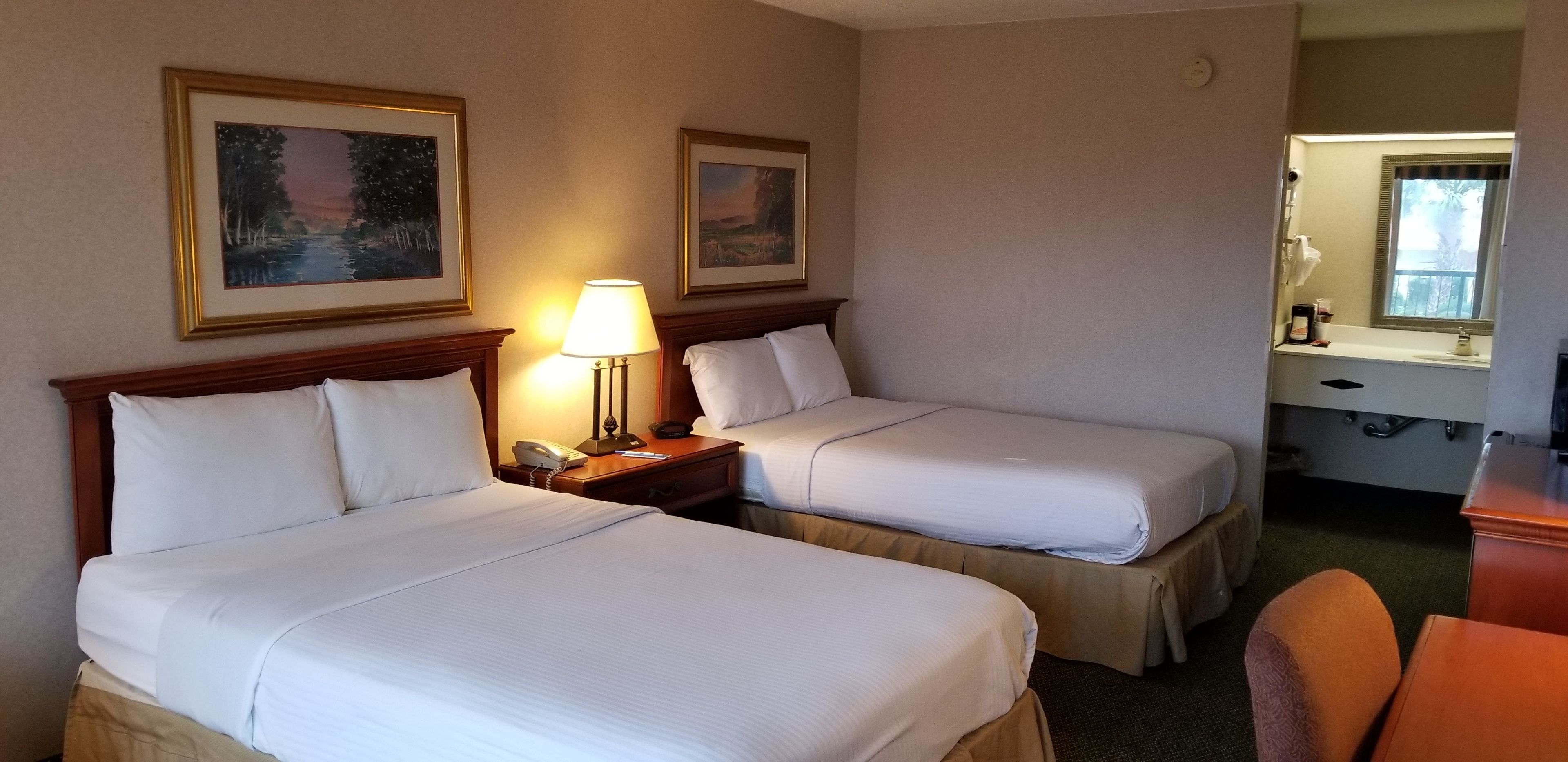 A hotel room with two beds, a nightstand with a lamp, a small table with a coffee maker, and a mirror.