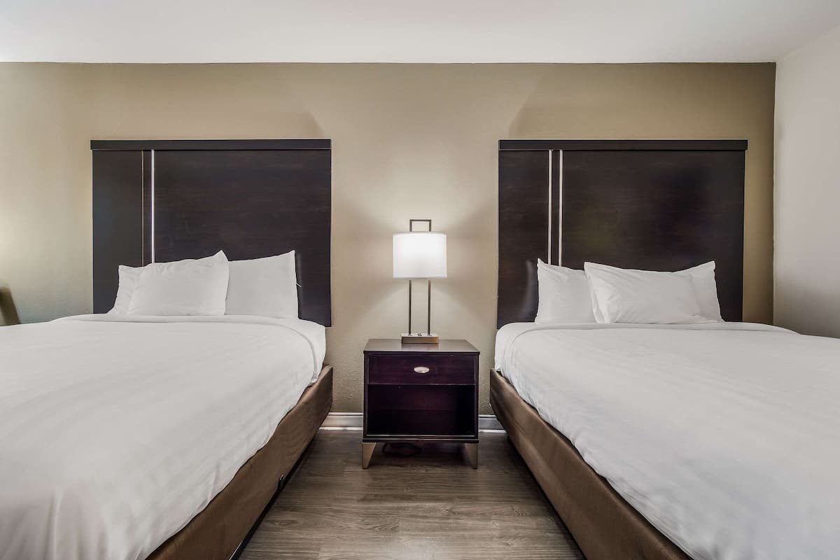 Quarto de hotel com duas camas, uma mesa de cabeceira com abajur e piso de madeira.