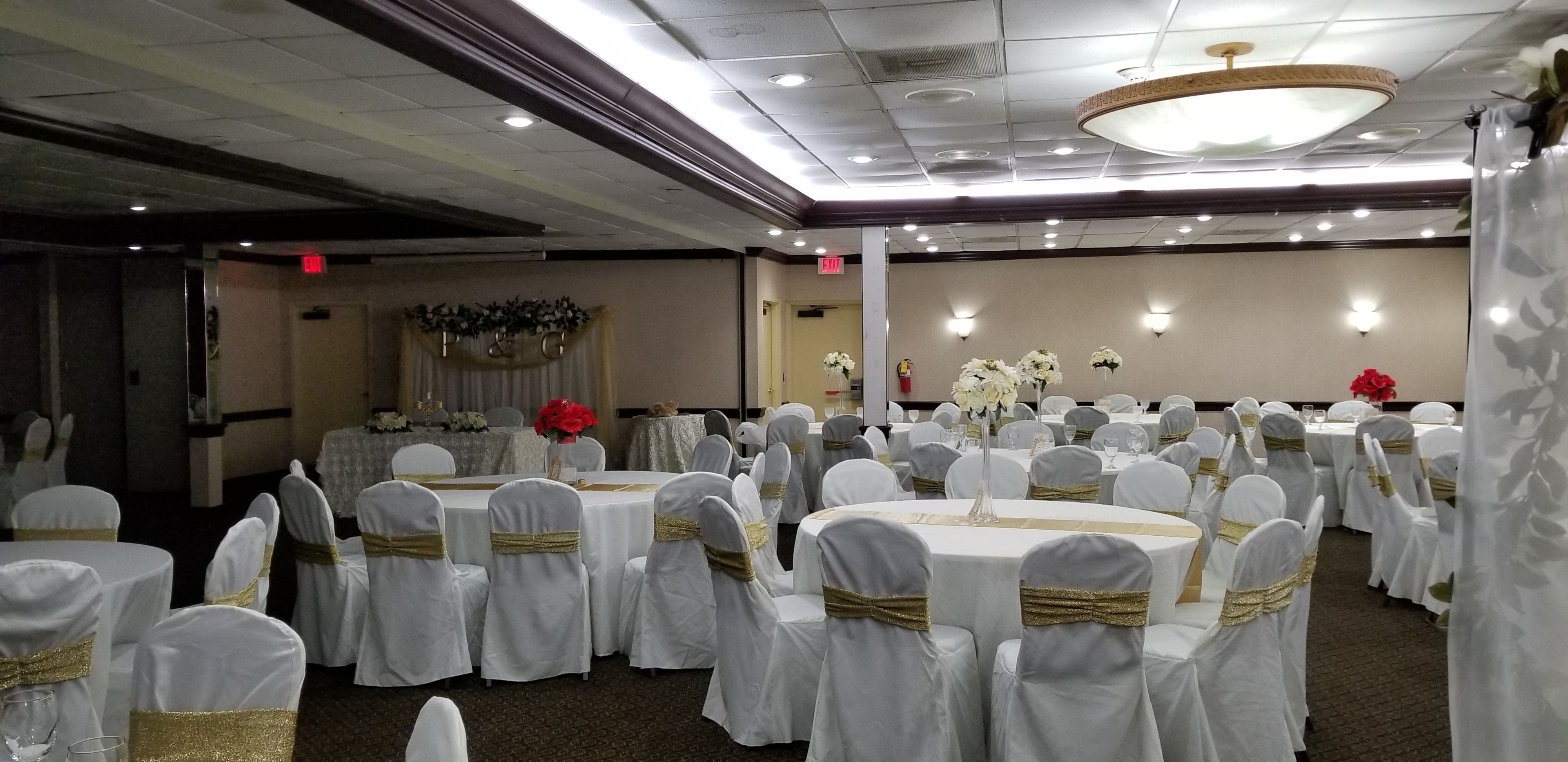 Un salón de banquetes con mesas redondas, sillas blancas con cintas doradas y arreglos florales.
