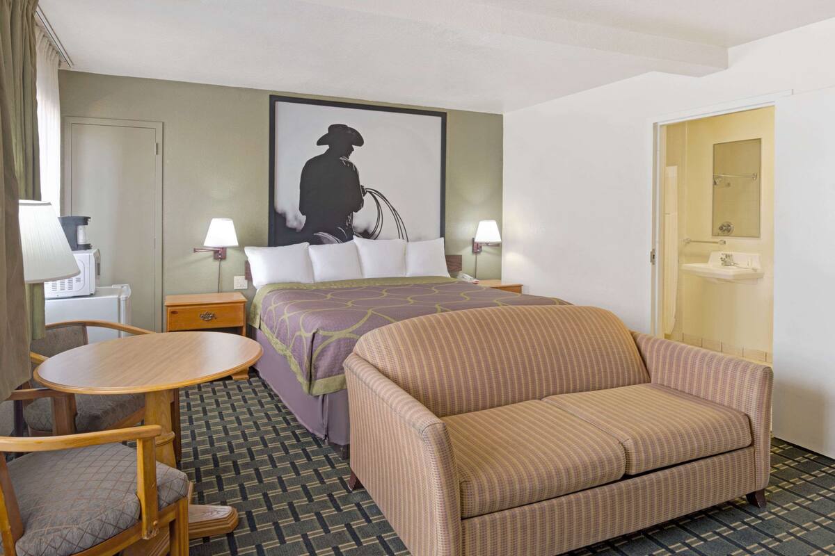 A hotel room with a bed, a sofa, a small table, two chairs, a desk, a lamp, a painting, and a bathroom door.