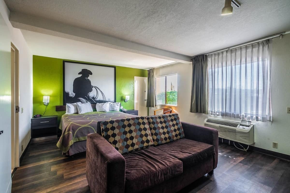 A hotel room with a bed, a sofa, a window with curtains, a wall-mounted air conditioner, and a painting on the wall.