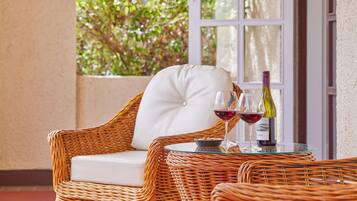 Une chaise en osier avec un coussin blanc, une table en verre avec une bouteille de vin et un verre, et une vue sur la verdure par une fenêtre.