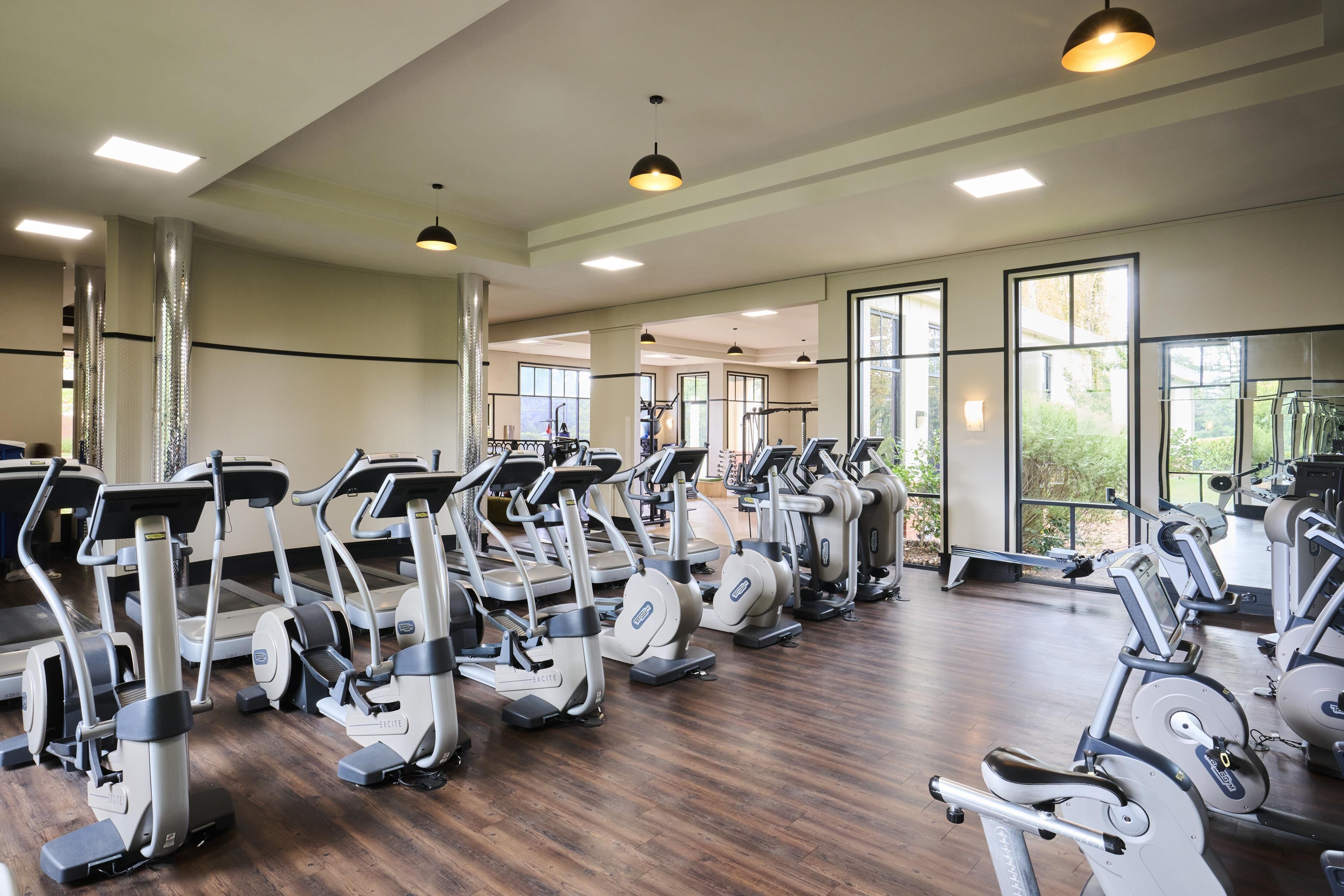 A gym with multiple treadmills and exercise equipment, large windows, and a wooden floor.