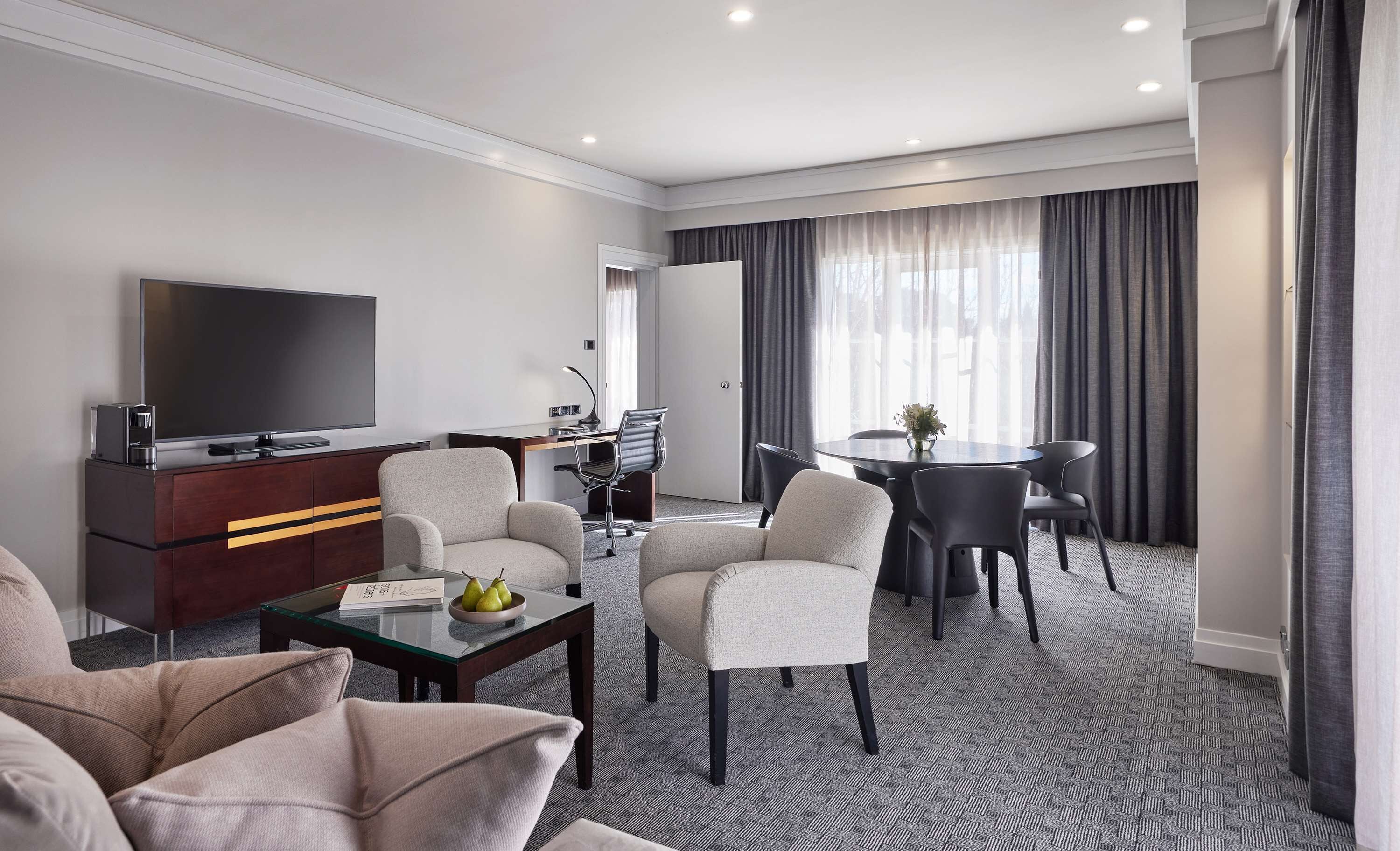 A modern hotel room with a flat-screen TV, a sofa, a dining table with chairs, and a small desk with a chair.