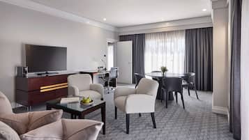 A modern hotel room with a flat-screen TV, a sofa, a dining table with chairs, and a small desk with a chair.