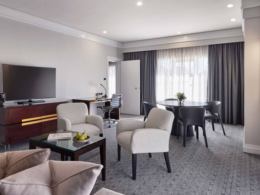 A modern hotel room with a flat-screen TV, a sofa, a dining table with chairs, and a small desk with a chair.