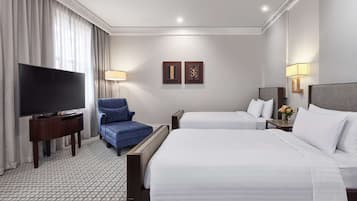 A hotel room with two beds, a TV, a blue armchair, and a small table with a lamp.