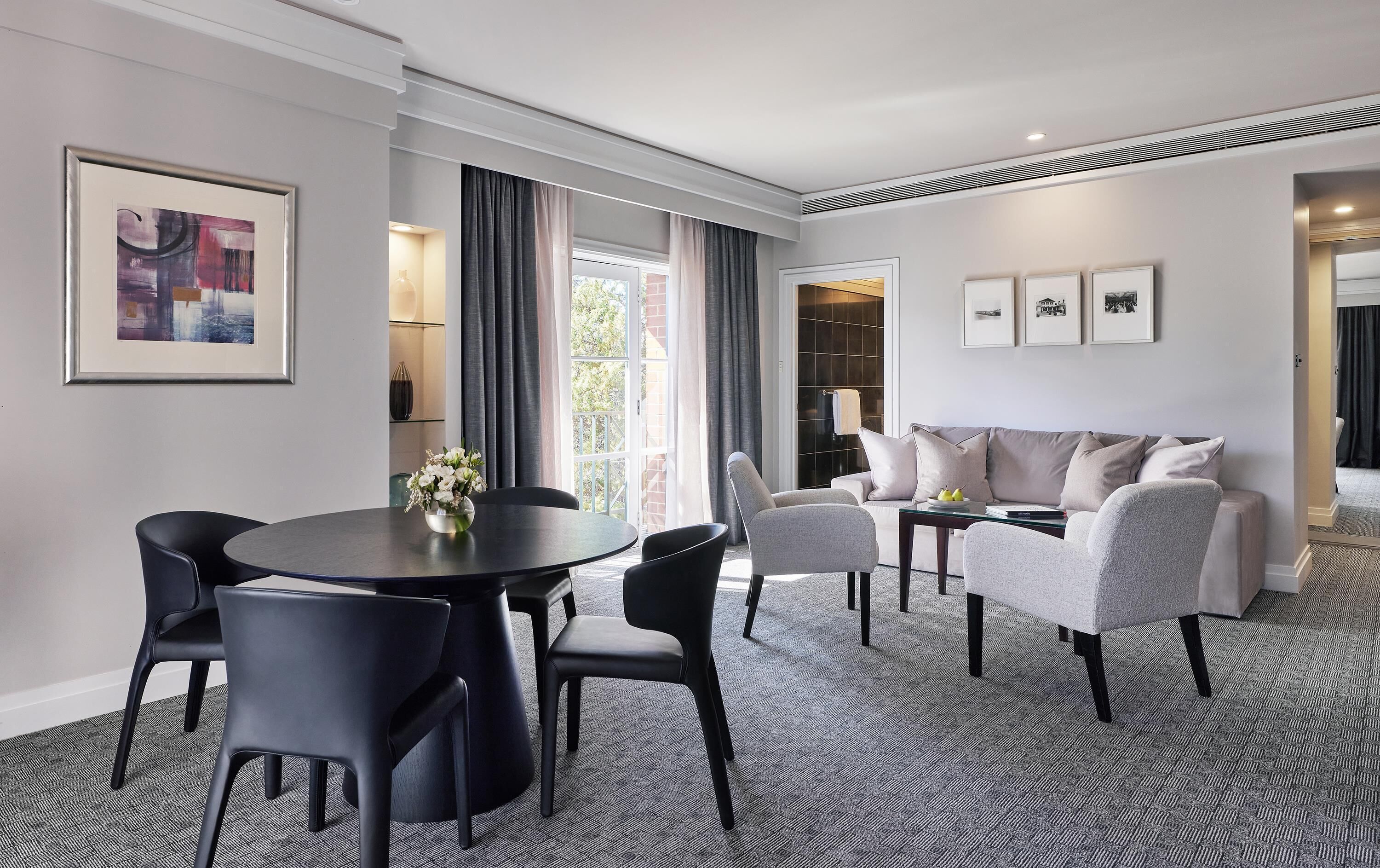 A modern hotel room with a dining area, a sofa set, and a large window with curtains.