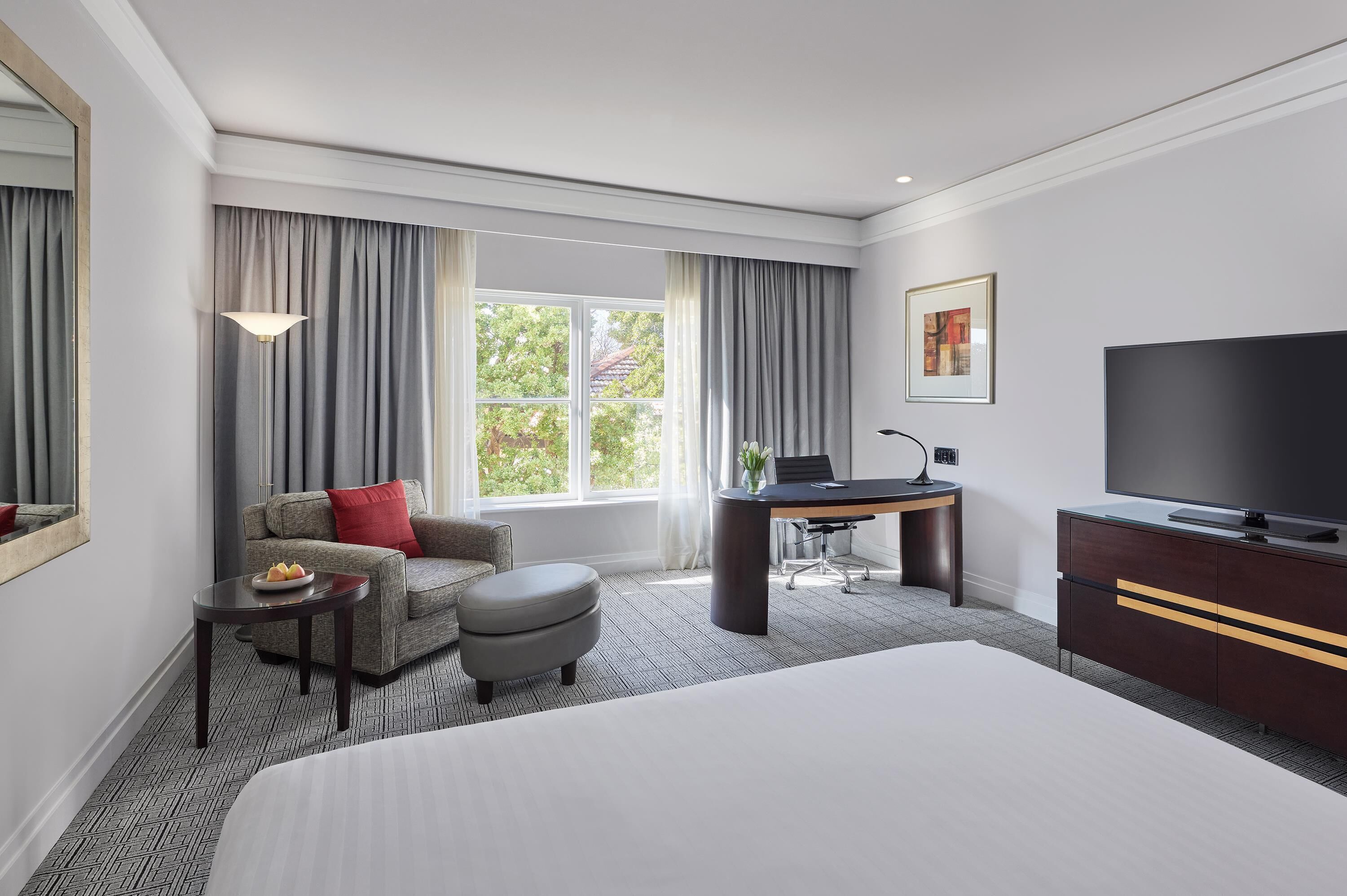 A hotel room with a large bed, a desk with a chair, a sofa with a red pillow, a small table with a fruit bowl, and a TV on a cabinet.