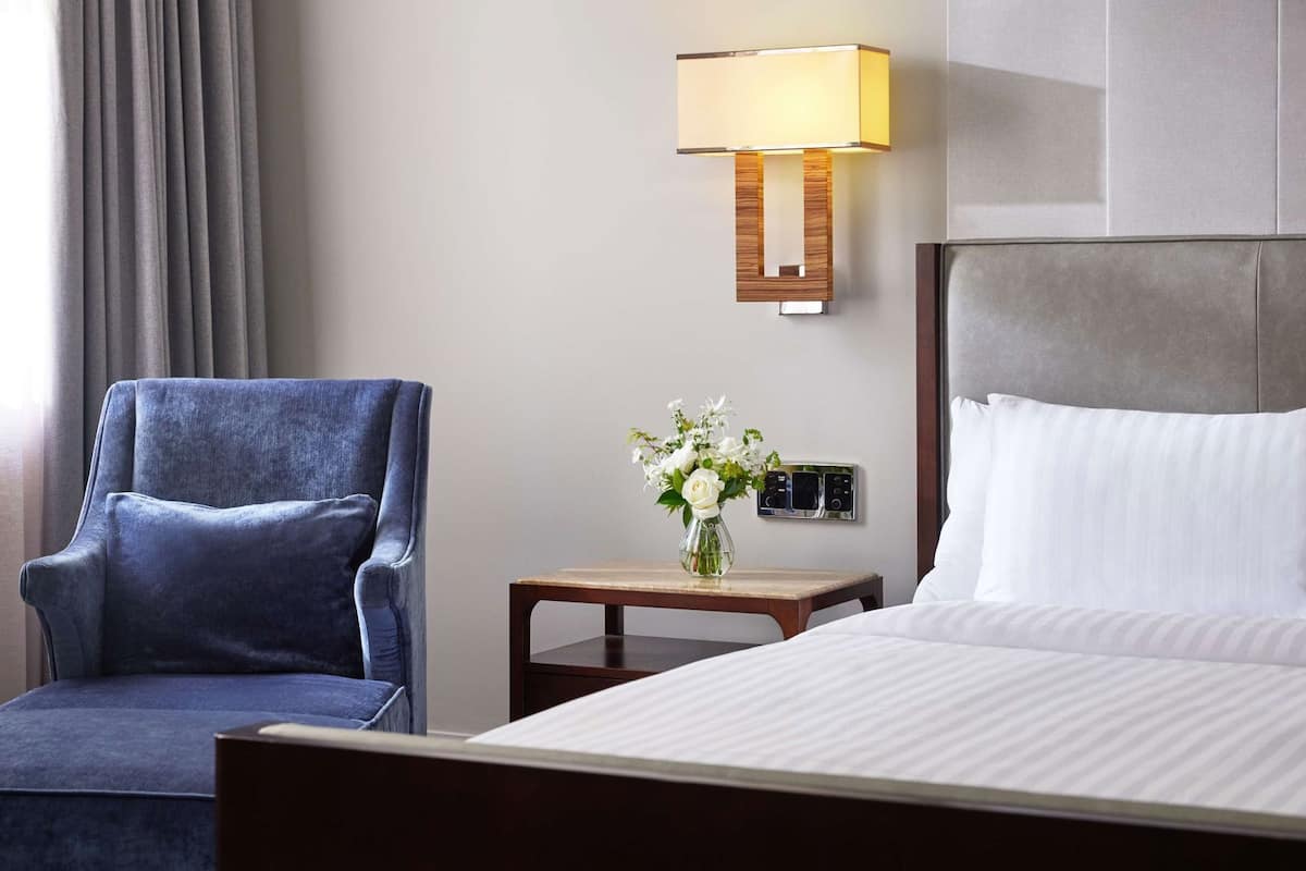 Een hotelkamer met een bed, een blauwe fauteuil, een nachtkastje met een lamp en een bedieningspaneel aan de muur.
