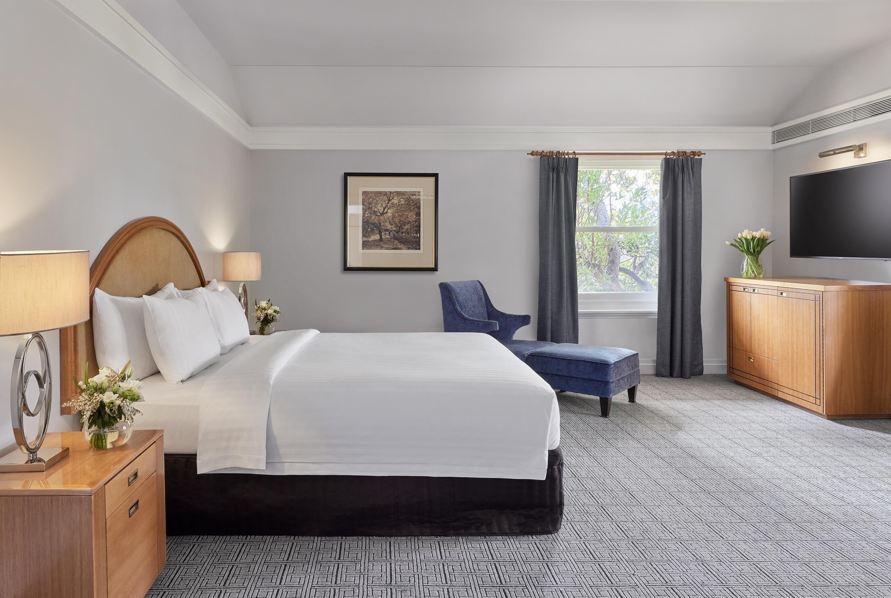 A hotel room with a large bed, a desk with a lamp, a chair, a TV, and a window with curtains.