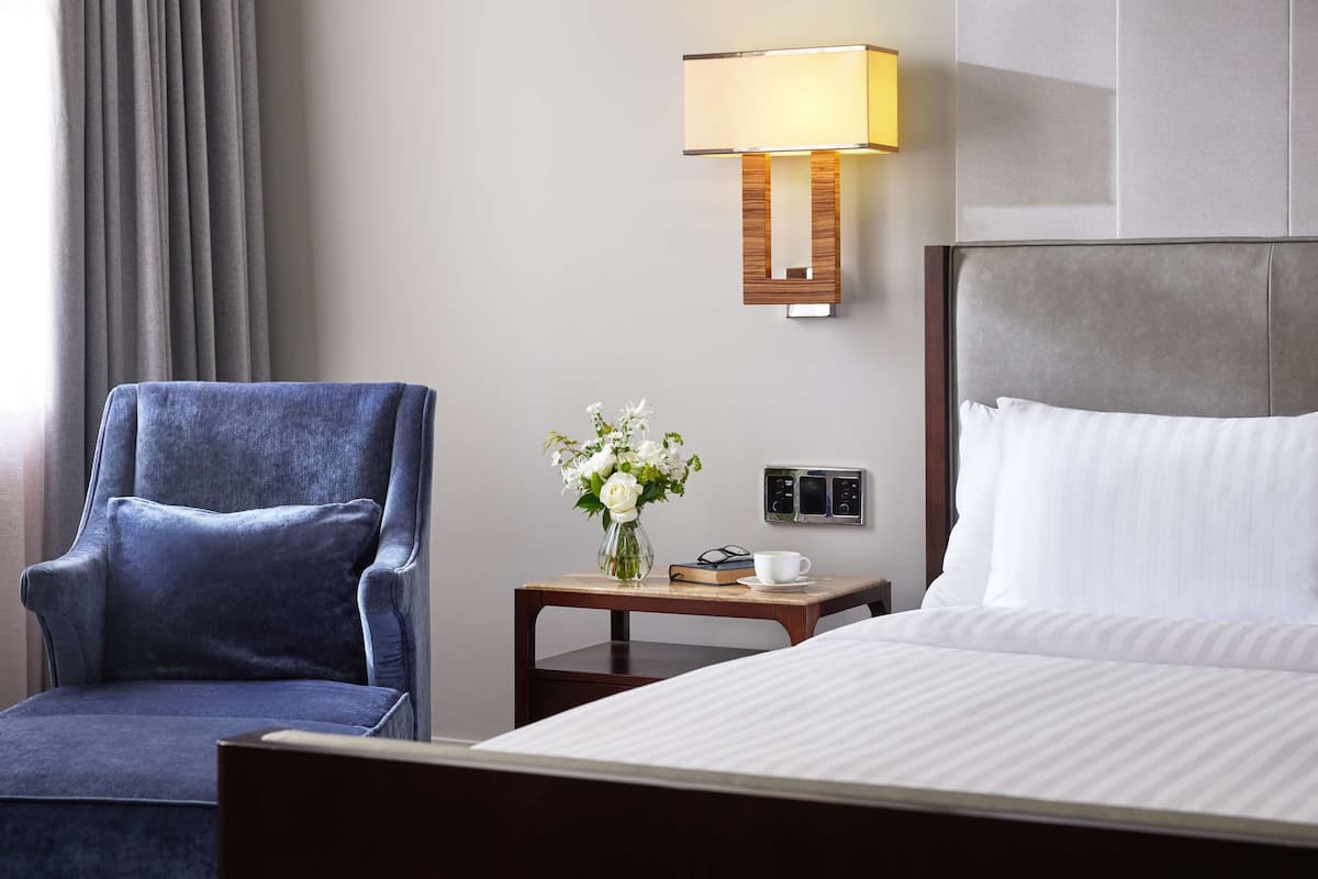 Een hotelkamer met een bed, een blauwe fauteuil, een nachtkastje met een lamp, een vaas met bloemen en een klein klokje.