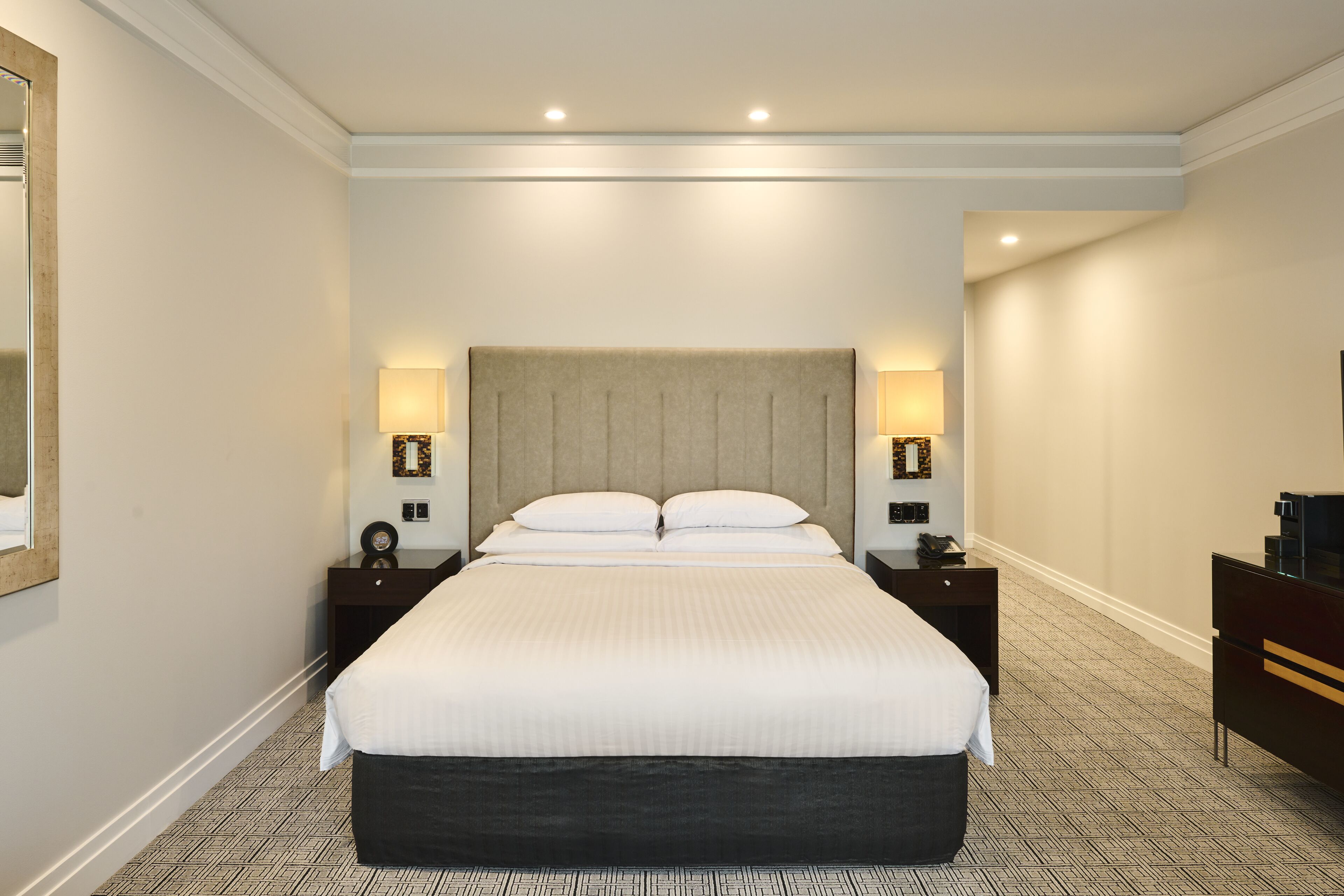 Une chambre d’hôtel avec un grand lit, deux tables de chevet, une petite table, une commode et un miroir sur le mur de gauche.