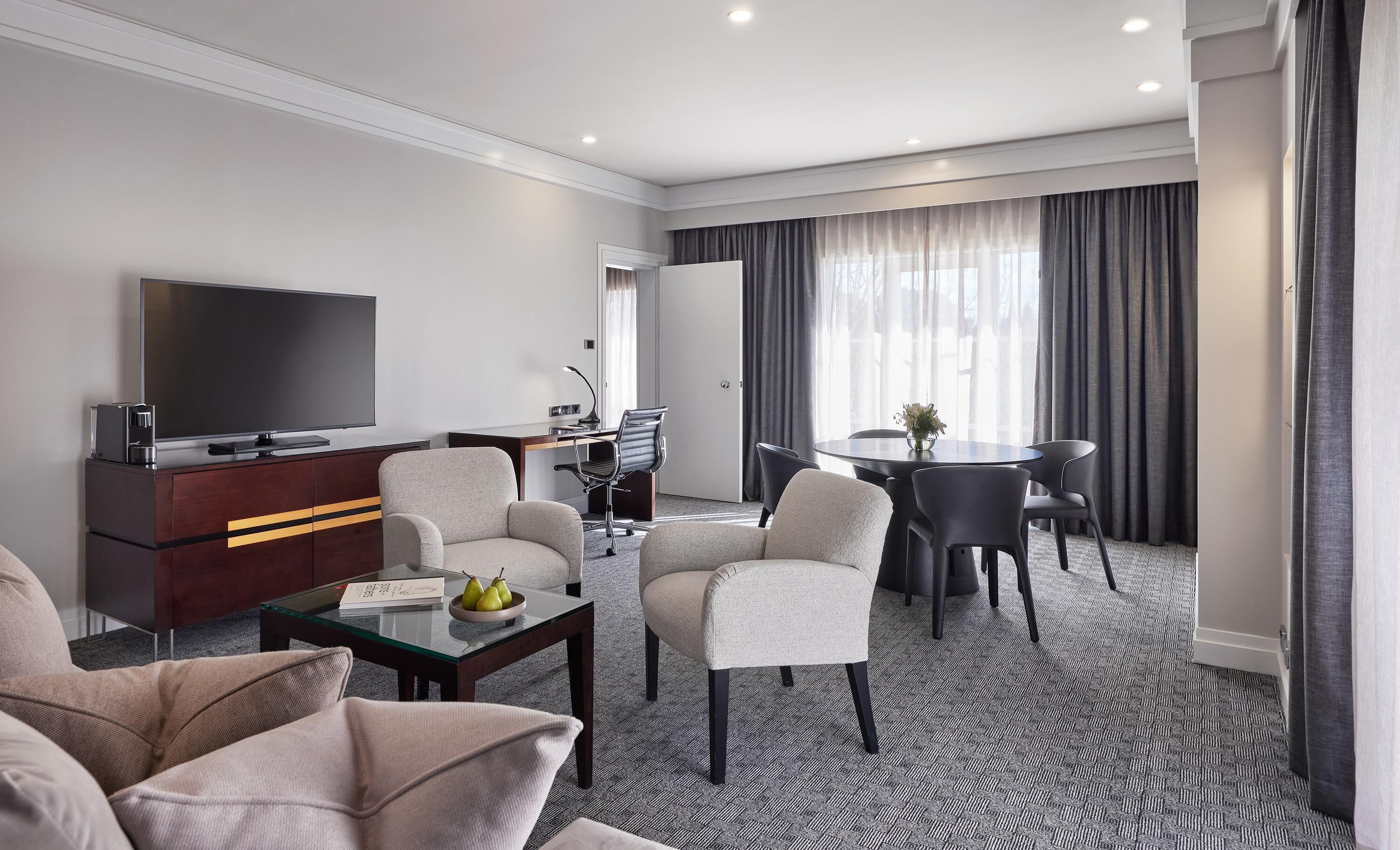 Ein modernes Hotelzimmer mit Flachbildfernseher, einer Sitzecke, einem Esstisch mit Stühlen, einem kleinen Schreibtisch inklusive Stuhl und einem großen Fenster mit Vorhängen.