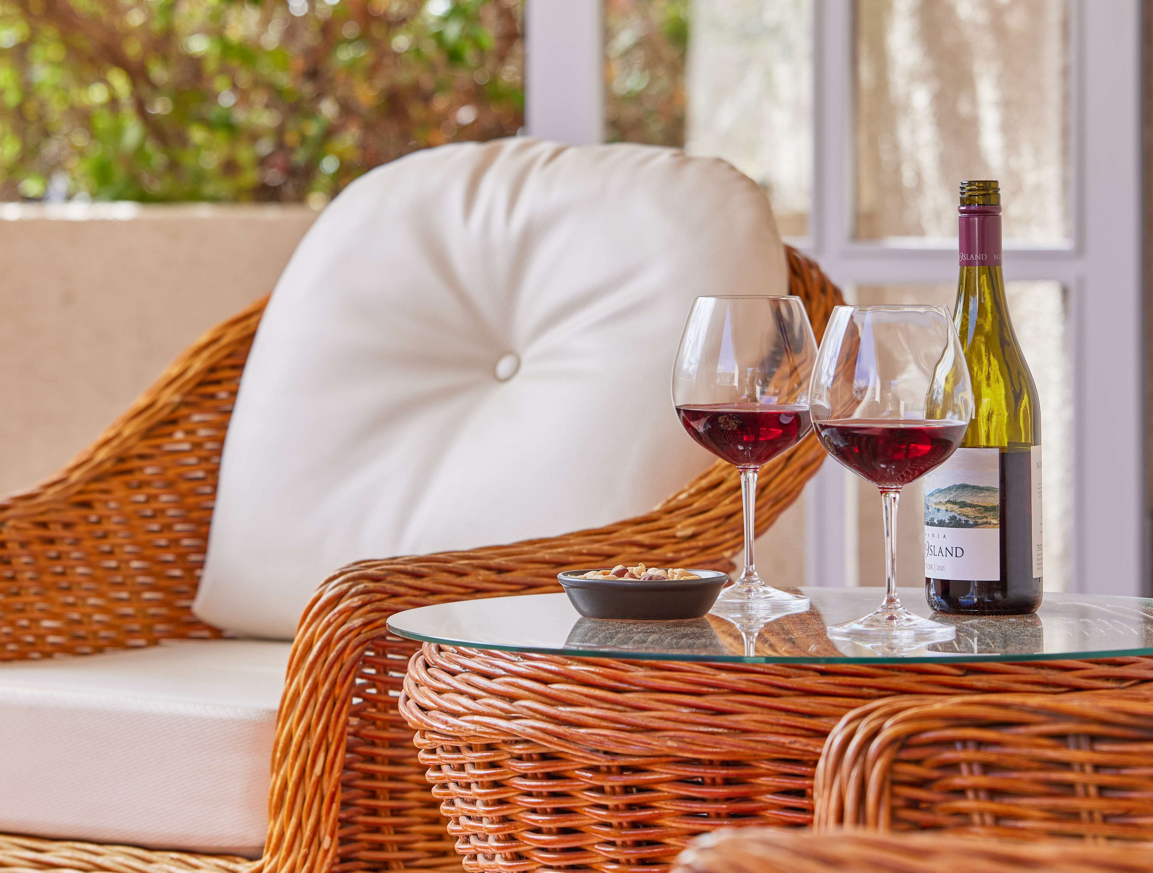 A wicker chair with a cushion, a glass table with two wine glasses and a bottle, and a bowl of nuts.