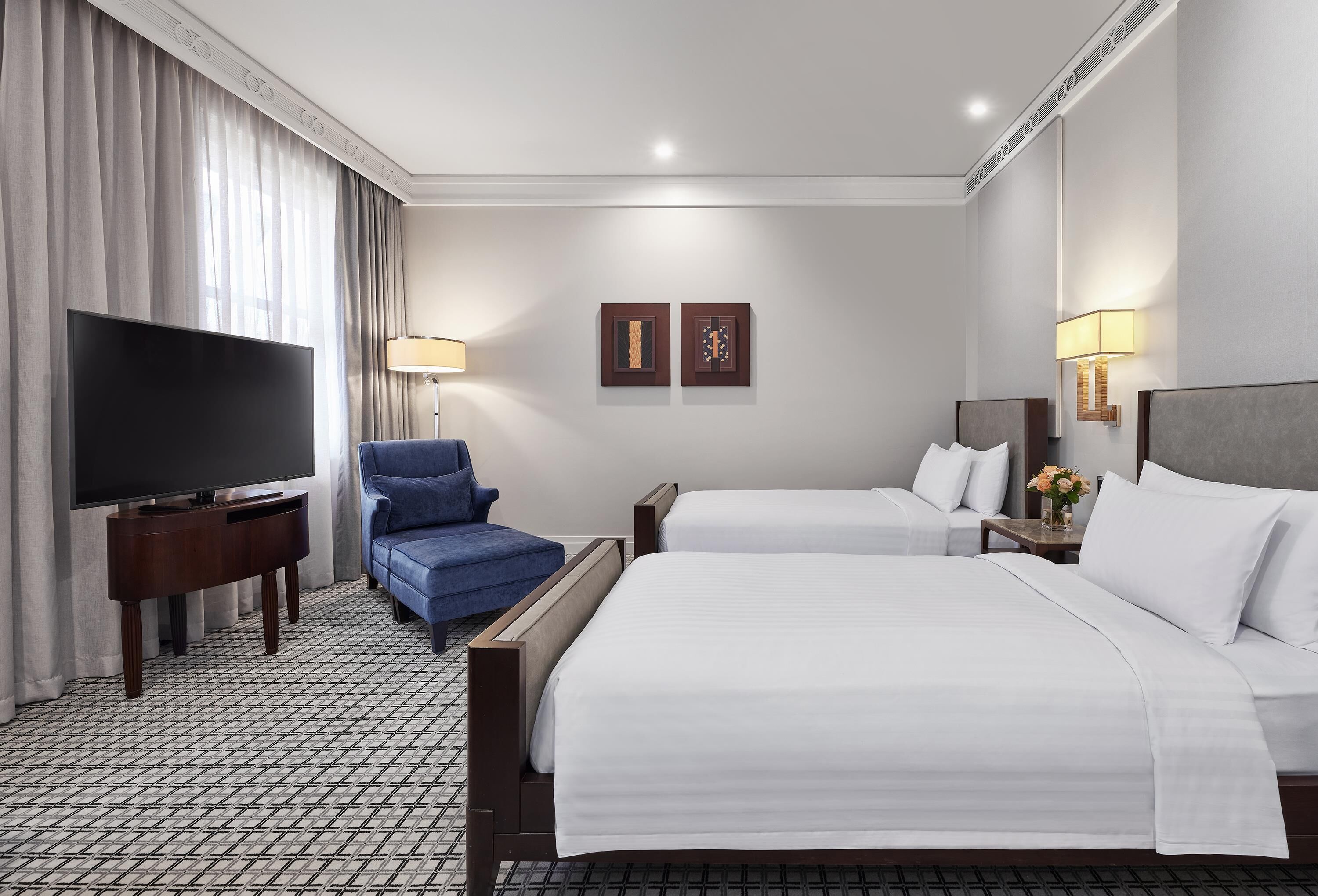 Habitación de hotel con dos camas, un televisor de pantalla plana, un sillón azul y dos lámparas de pared.